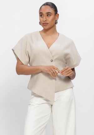 Kobieta z ciemnymi włosami upiętymi w koka, ubrana w beżową bluzkę z krótkim rękawem o kroju kopertowym oraz białe spodnie, patrząca na bok, z dłońmi lekko złączonymi.