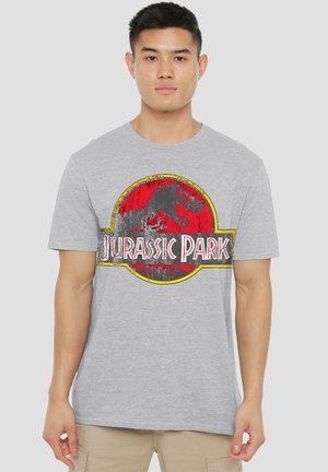 Young man wearing light gray T-shirt with red and black Jurassic Park logo standing against plain background.