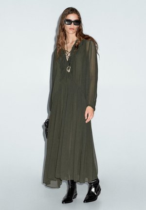 Robe longue légère de couleur vert olive avec des détails froncés et un décolleté en V. Associée à des bottines noires brillantes et des lunettes de soleil oversize noires.