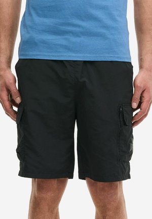 Hombre con pantalones cortos cargo negros con bolsillos laterales y camiseta azul, de pie con los brazos relajados a los lados.