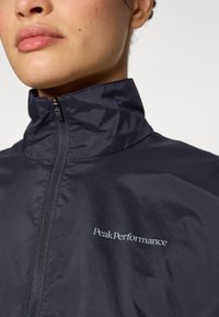 Marineblauwe, lichtgewicht jas met hoge kraag en volledige rits. Kenmerken zijn een logo, gladde textuur en een slank ontwerp.