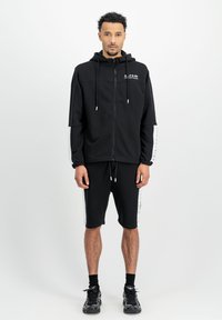 Hoodie zippé noir avec cordons de serrage et détails graphiques blancs, associé à un short noir avec des accents latéraux blancs, porté avec des baskets noires.