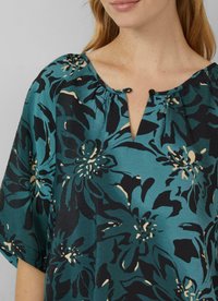 Top floral en bleu sarcelle avec des motifs noirs et crème. Comprend une patte de boutonnage avec deux boutons, un col froncé et des manches courtes et amples.