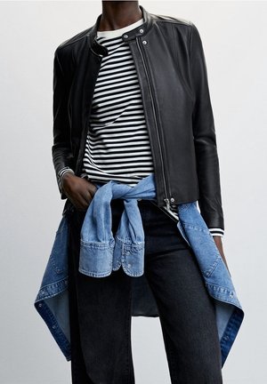 Person iført sort læderjakke over sort-hvidstribet skjorte med en blå denimjakke bundet om livet og sorte bukser.