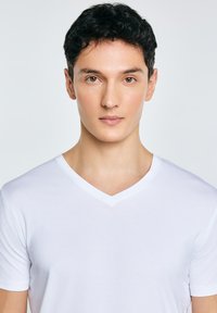 Joven con cabello corto y negro, vistiendo una camiseta blanca de cuello en V, mirando hacia adelante sobre un fondo liso de color gris claro.