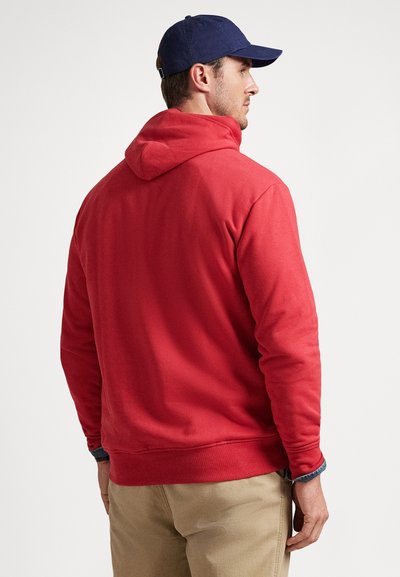 Άνδρας που φοράει κόκκινο φούτερ με κουκούλα, μπεζ παντελόνι και μπλε σκούρο καπέλο μπέιζμπολ, σ�τέκεται πλάτη σε λευκό φόντο.