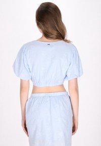 Vaaleansininen lyhythihainen crop-top, jossa on puffihihat, kukkakuosiset tekstiilit, joustava helma ja pieni metallinen yksityiskohta takana.