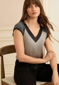 Graue Bluse mit V-Ausschnitt, schwarzem Saum und schimmernden schwarzen Ärmeln. Kombiniert mit schwarzen Hosen und sitzt auf einem Holzstuhl.