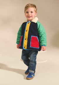Veste colorée pour enfants avec un corps en polaire bleu marine, des manches matelassées vertes, une fermeture éclair jaune à l'avant et des poches rouges. Portée avec un jean en denim et des chaussures bleues.