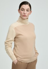 Frau mit dunklem Haar, die einen beigen und cremfarbenen Rollkragenpullover und braune Hosen trägt, mit den Händen in den Taschen, blickt zur Seite.