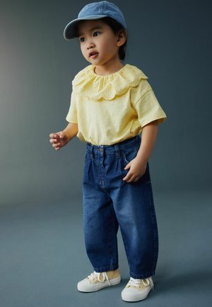 Väikelaps, kes kannab kollast pluusi pitskaelusega, siniseid teksaseid, beežid tossud ja sinist nokamütsi, seisab tavalise halli tausta ees.