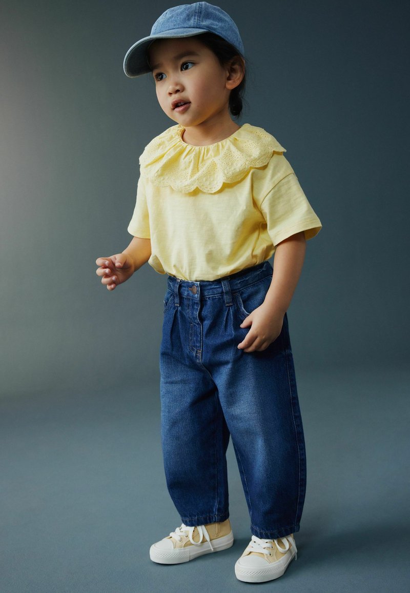 Tout-petit portant une chemise jaune avec un col en dentelle, un jean bleu, des baskets beige et une casquette bleue, debout devant un fond gris uni.