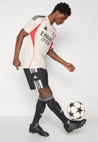 Vyrų futbolo apranga, sudaryta iš šviesiai pilkos marškinėlių su raudonais akcentais, juodų šortų su baltais raštais, juodų kojinių ir juodų lauko batelių, mušant futbolo kamuolį.