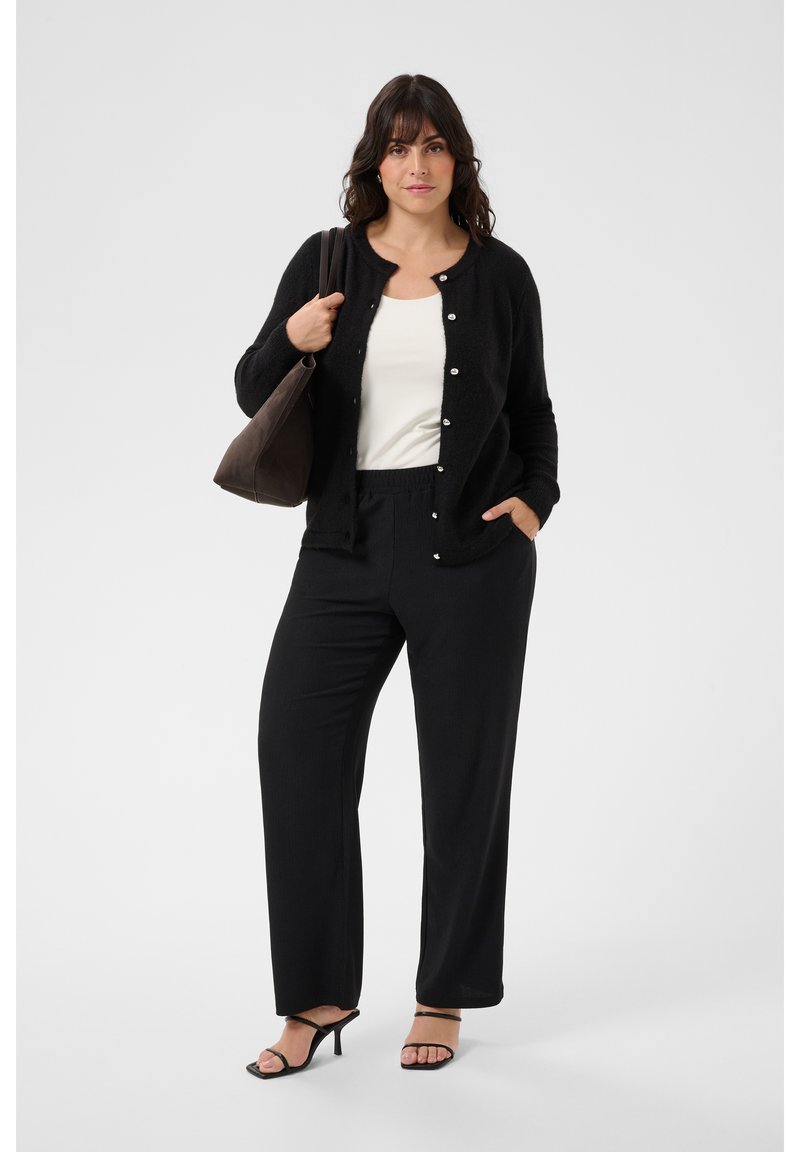 Femme portant un cardigan noir, un haut blanc et un pantalon noir, tenant un sac marron avec une main dans la poche, debout sur fond blanc.