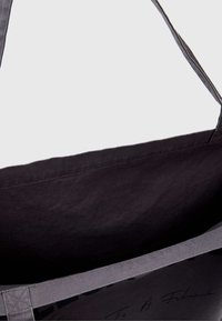 Tamsiai pilkas drobinis maišas, turintis paprastą dizainą su tvirtais dirželiais. Vidus išklotas atitinkančios tamsios spalvos audiniu su ranka rašytos parašo detale.