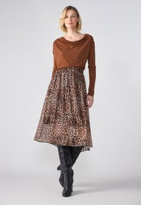 Ein braunes, langärmliges Oberteil mit Drapierung, kombiniert mit einem gestuften Rock im Leopardenmuster. Schwarze kniehohe Stiefel runden den Look ab. Glatte Texturen überall.