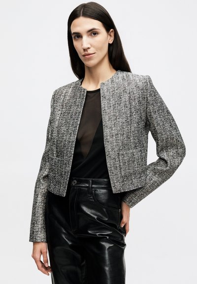 Žena s rovnými tmavými vlasy nosí texturovanou šedou krátkou bundu přes černý průsvitný top a černé kožené kalhoty, stojící na bílém pozadí.