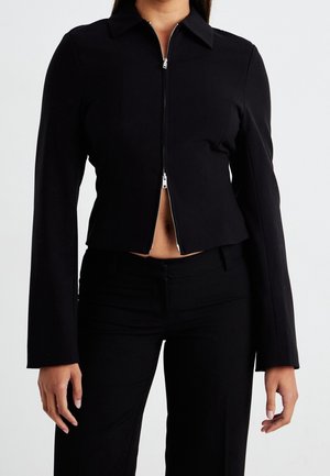 Woman wearing a fitted black long-sleeve zip-up jacket with a collar and matching black pants against a plain background.