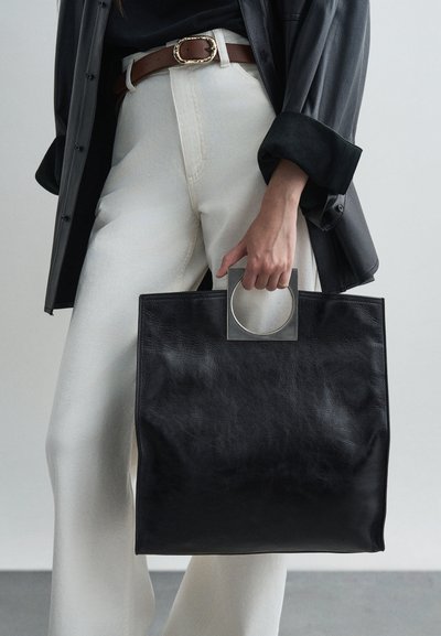 Black leather tote bag with a square metallic handle. The bag has a smooth texture and structured shape, paired with cream-colored trousers.