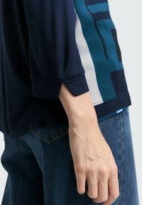 Marinblå topp med vida ärmar och sprund, kombinerad med jeans i denim. Toppen har en geometrisk design i blått och grått på sidan.