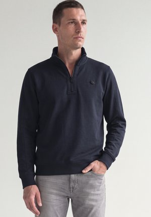 Man wearing a black quarter-zip sweatshirt and light gray jeans, standing with one hand in pocket, looking to the side on a plain background.