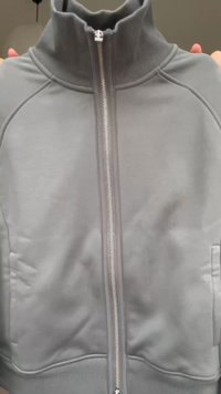 Giacca grigia chiaro con zip e collo alto a coste, tenuta con le mani su uno sfondo neutro.