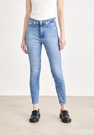Kvinde iført lyseblå højtaljede skinny jeans og sorte loafers, stående mod en ensfarvet hvid baggrund.