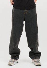 Mustat farkut leveällä lahkeella, joissa on haalistunut pinta, varustettuna perinteisellä napilla ja vetoketjulla, yhdistettynä vaaleisiin lenkkareihin.