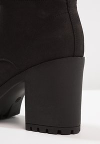 Black suede heeled boot featuring a textured platform, rounded heel, and stitching details along the seams. Solid rubber sole with grooves.
