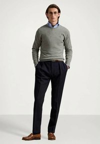 Man som bär en grå tröja, blå skjorta med krage, marinblå ränder på byxorna, bruna loafers och runda glasögon, står med händerna i fickorna.