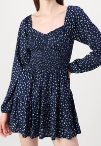 Mörkblå blommig klänning med långa ärmar, smockad bodice och volangkant. Har vita blommönster över hela klänningen och en fyrkantig halsringning.