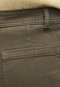 Κοντινή άποψη της πίσω τσέπης και της θηλιάς της ζώνης σε ελιάς πράσινο παντελόνι με πουλόβερ πλεκτό εκρού πάνω από τη μέση.