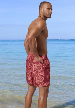 Muskulöser Mann in rot gemusterten Badeshorts steht im flachen, klaren Ozeanwasser und blickt über die Schulter gegen einen blauen Himmel.