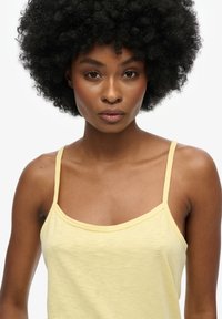 Top de tirantes finos en algodón amarillo, con escote redondeado y una textura suave y ligera. Costuras visibles cerca del escote.