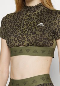 Top corto de manga corta en verde olivo con estampado de leopardo negro. Presenta un cuello alto y una banda elástica con el logo de Adidas.