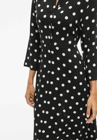 Šaty s knoflíkovým zapínáním, vyrobené z černé látky s velkými bílými puntíky, dlouhé rukávy a úzký pas. Hladká textura s klasickým designem.