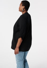Zwarte top met lange mouwen en opgerolde mouwen, ontspannen pasvorm en een subtiel textuurpatroon, gecombineerd met lichtblauwe jeans.