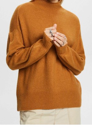 Persoon die een roestkleurige wollen trui met opgerolde manchetten draagt, handen gevouwen met meerdere zilveren ringen, gecombineerd met lichtbeige broek.