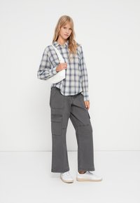 Camisa xadrez azul e branca de mangas longas, acompanhada de calças cargo cinzentas com bolsos laterais e ténis brancos.