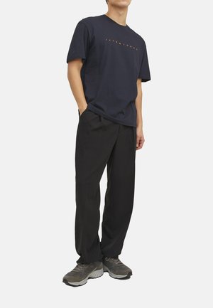 Marineblaues T-Shirt mit orangefarbenem "JACK & JONES"-Logo, kombiniert mit schwarzen locker geschnittenen Hosen und grauen Sportschuhen. Glatter Stoff und entspannte Silhouette.