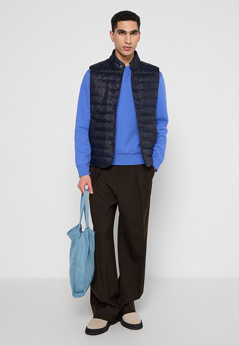 Granatowa kamizelka puchowa na tle niebieskiego swetra, w połączeniu z brązowymi spodniami o szerokich nogawkach i beżowymi butami. Trzyma jasnoniebieską torbę tote.