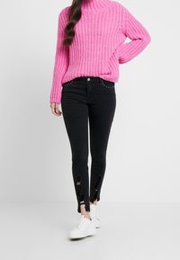 Suéter grueso rosa con cuello alto, combinado con jeans ajustados negros con rodillas desgastadas y detalles de tachuelas plateadas, usado con zapatillas blancas.
