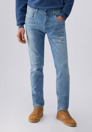 Muž v světle modrých opotřebovaných džínách, modré mikině a béžových semišových teniskách, stojící s jednou rukou v kapse před jednobarevným pozadím.