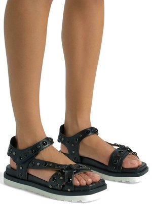 Black leather sandals feature cut-out design with circular studs, adjustable straps, and a textured white sole, showing toes.