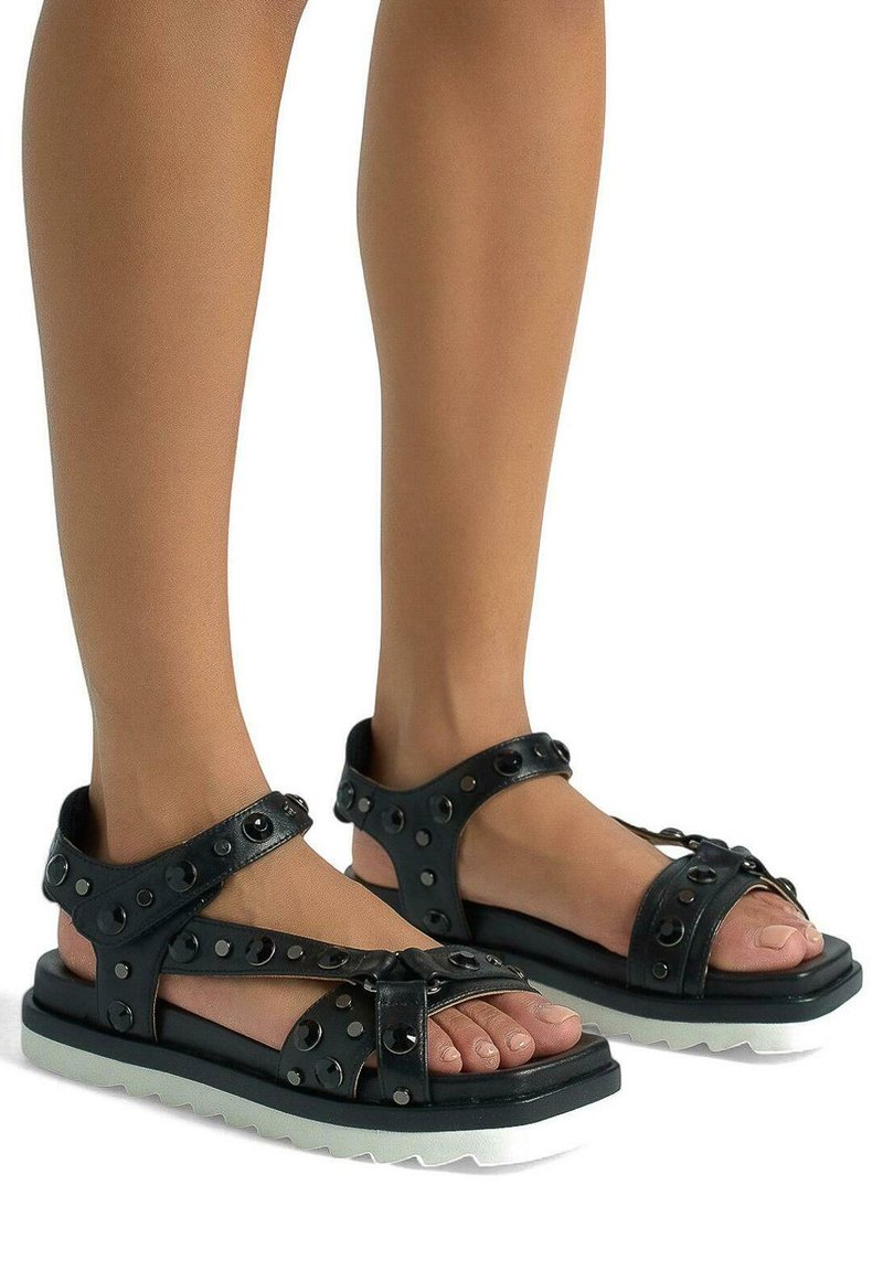 Las sandalias de cuero negro cuentan con un diseño recortado con clavos circulares, correas ajustables y una suela blanca texturizada, que muestra los dedos de los pies.
