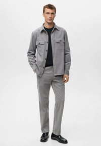 Veste-chemise en velours côtelé gris avec deux poches poitrine, associée à un pantalon texturé assorti. T-shirt noir à col rond en dessous. Chaussures noires.