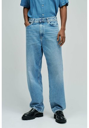 Persona con vaqueros sueltos de color azul claro, zapatos de cuero negros y una camisa azul de manga corta con botones, con una mano en el bolsillo.