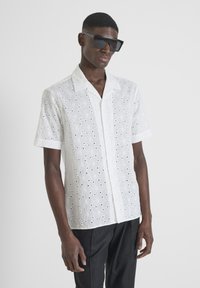 Korte mouwen witte shirt met patroon uitsparingen, voorzien van een klassieke kraag en knopen aan de voorkant. Gecombineerd met donkere broek en oversized zwarte zonnebril.