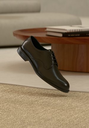 Black leather lace-up dress shoe floating above beige carpet with wooden coffee table and books in the background.