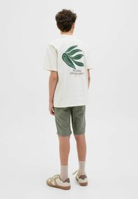 Off-white bomull t-shirt med grön abstrakt bladdesign och logotyp på baksidan, tillsammans med olivgröna shorts och beige sneakers.
